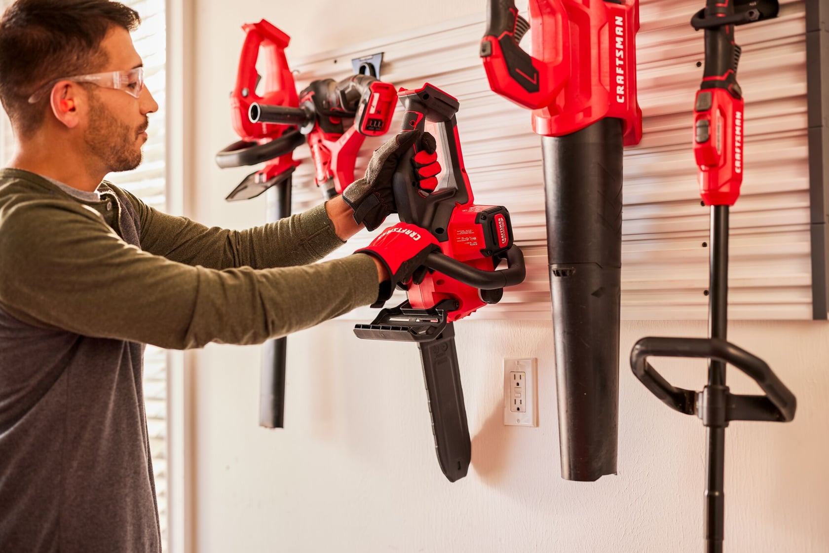 Photo of person hanging a Craftsman CMCCS630B cordless chainsaw on a wall-mounted tool organizer with other Craftsman tools.