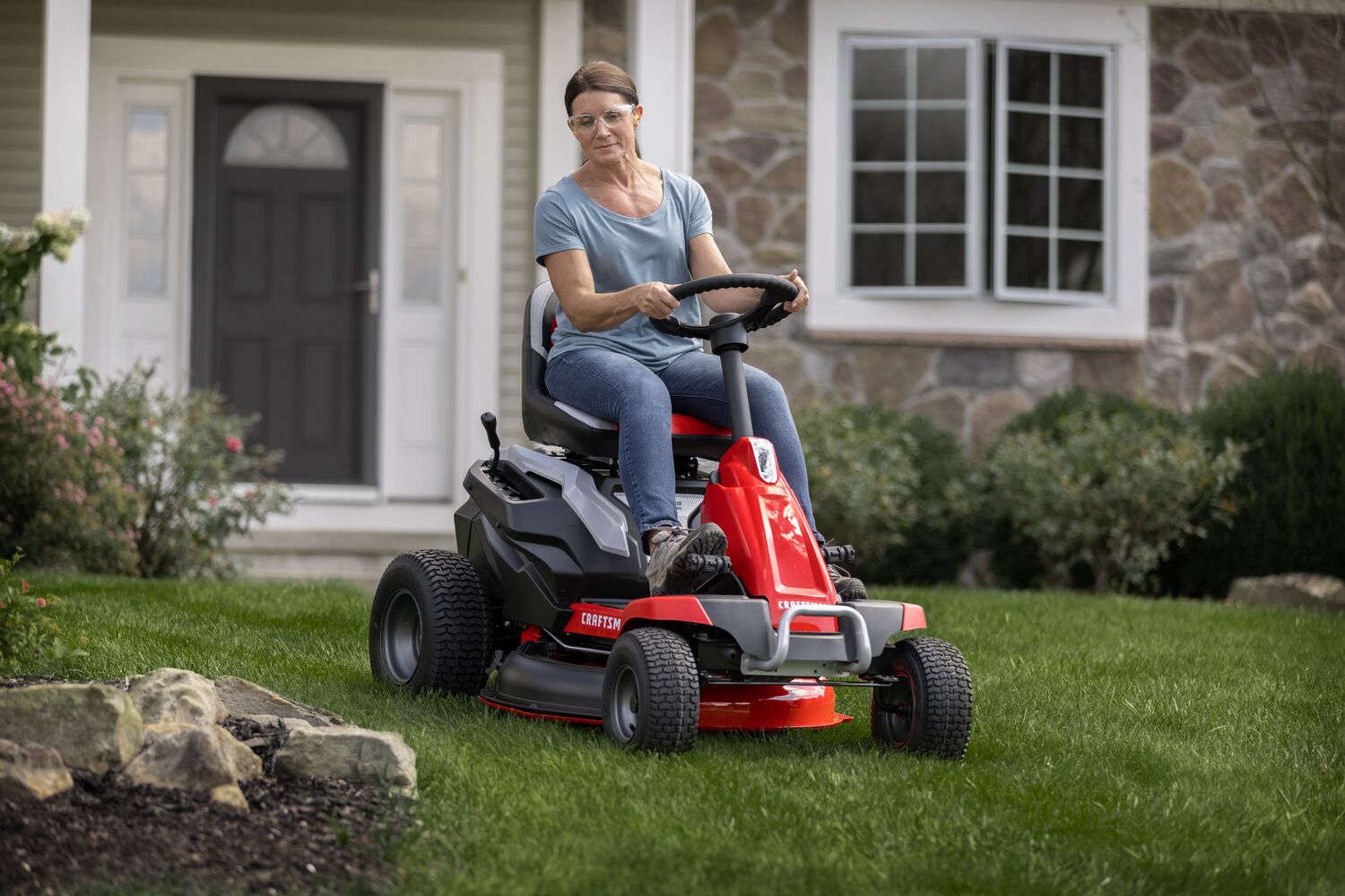 CRAFTSMAN Battery-Powered Compact Mower cutting grass around flowerbed with house in background.