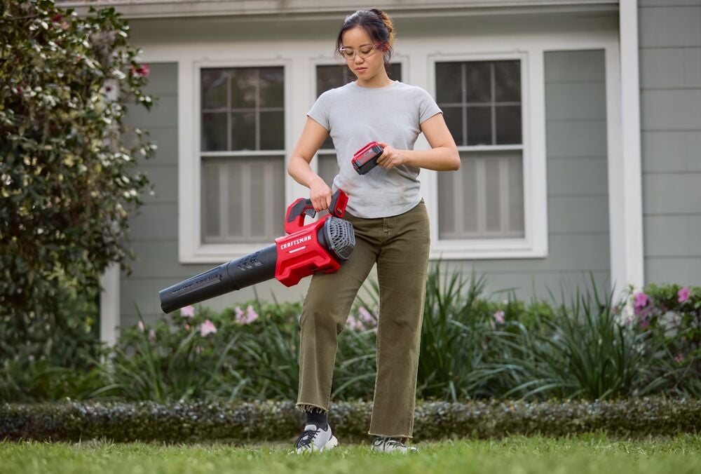Photo of person using Craftsman cordless leaf blower CMCBL730P1 in a yard, holding battery, with house in background.